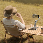 Person sitting in a field with a hat, using a smartphone on a stand at a small wooden table.